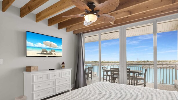 A bedroom with a large bed, a ceiling fan, a TV on the wall, a dresser, and a balcony with a view of the water.