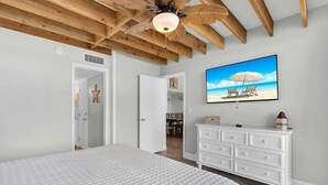 A bedroom with a large bed, wooden ceiling beams, a ceiling fan, a white dresser, and a beach-themed wall art.
