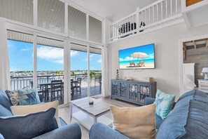 Un vaste salon avec un balcon donnant sur une marina, un téléviseur fixé au mur et un meuble moderne de couleur grise.
