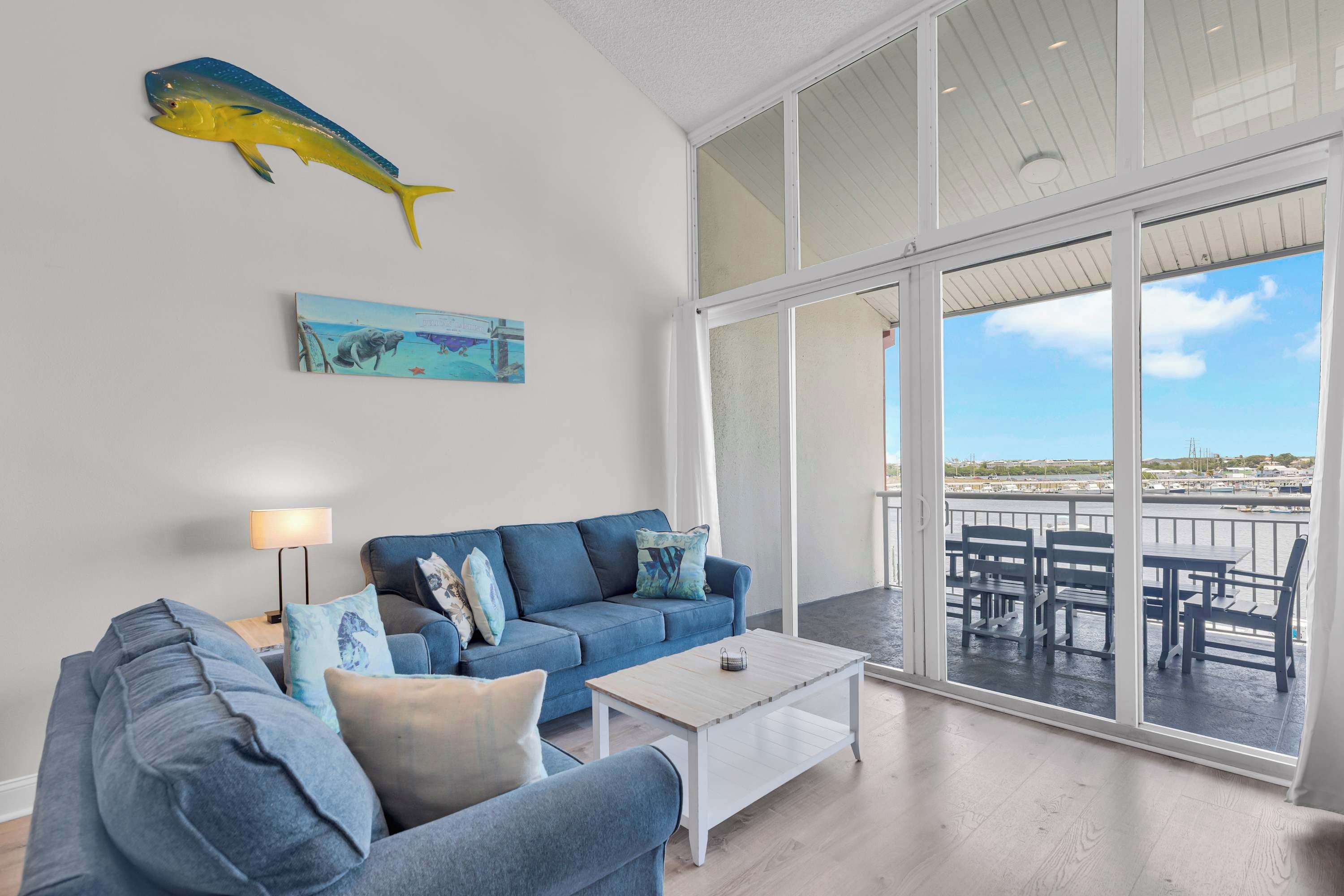 A modern living room with a blue sofa, a coffee table, and a large window overlooking a balcony with outdoor furniture.