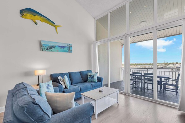 A modern living room with a blue sofa, a coffee table, and a large window overlooking a balcony with outdoor furniture.