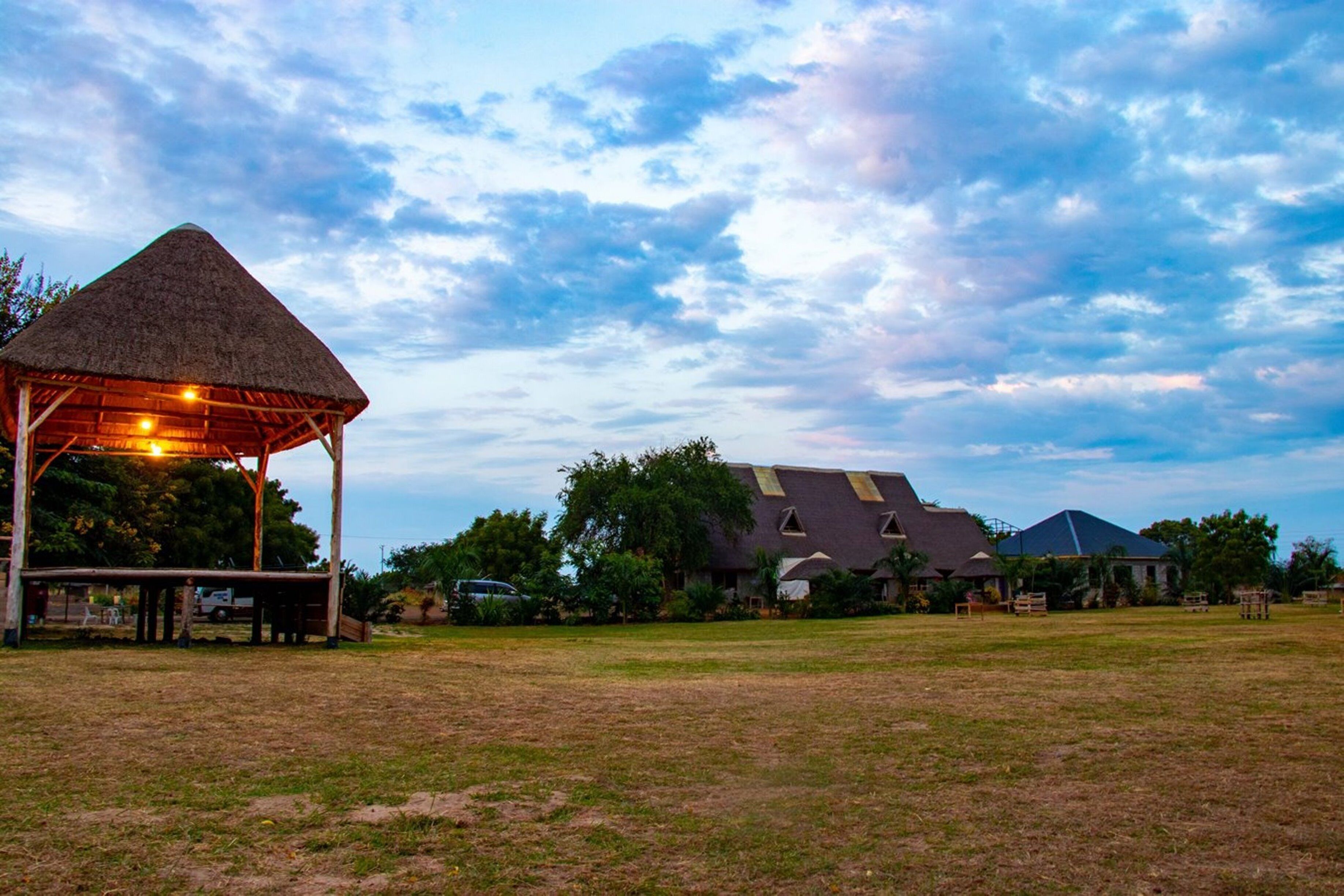 A thatched-roof pavilion with a wooden bench, surrounded by a grassy field and a large house with a sloped roof.