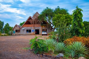 A rustic building with a brick facade and a thatched roof, surrounded by lush greenery and a gravel driveway. - Western Sands Hotel (Bulisa)