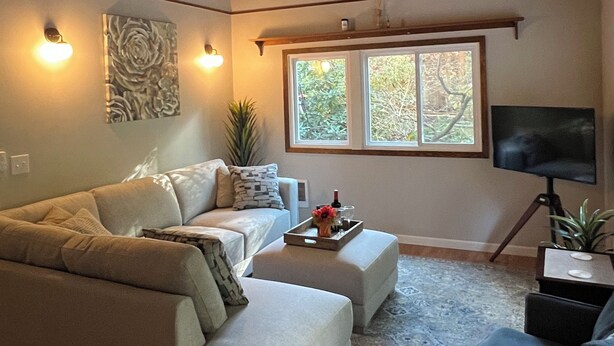 A living room with a sofa, ottoman, and a TV on a stand. - Charming Geneva Bungalow (Bellingham)