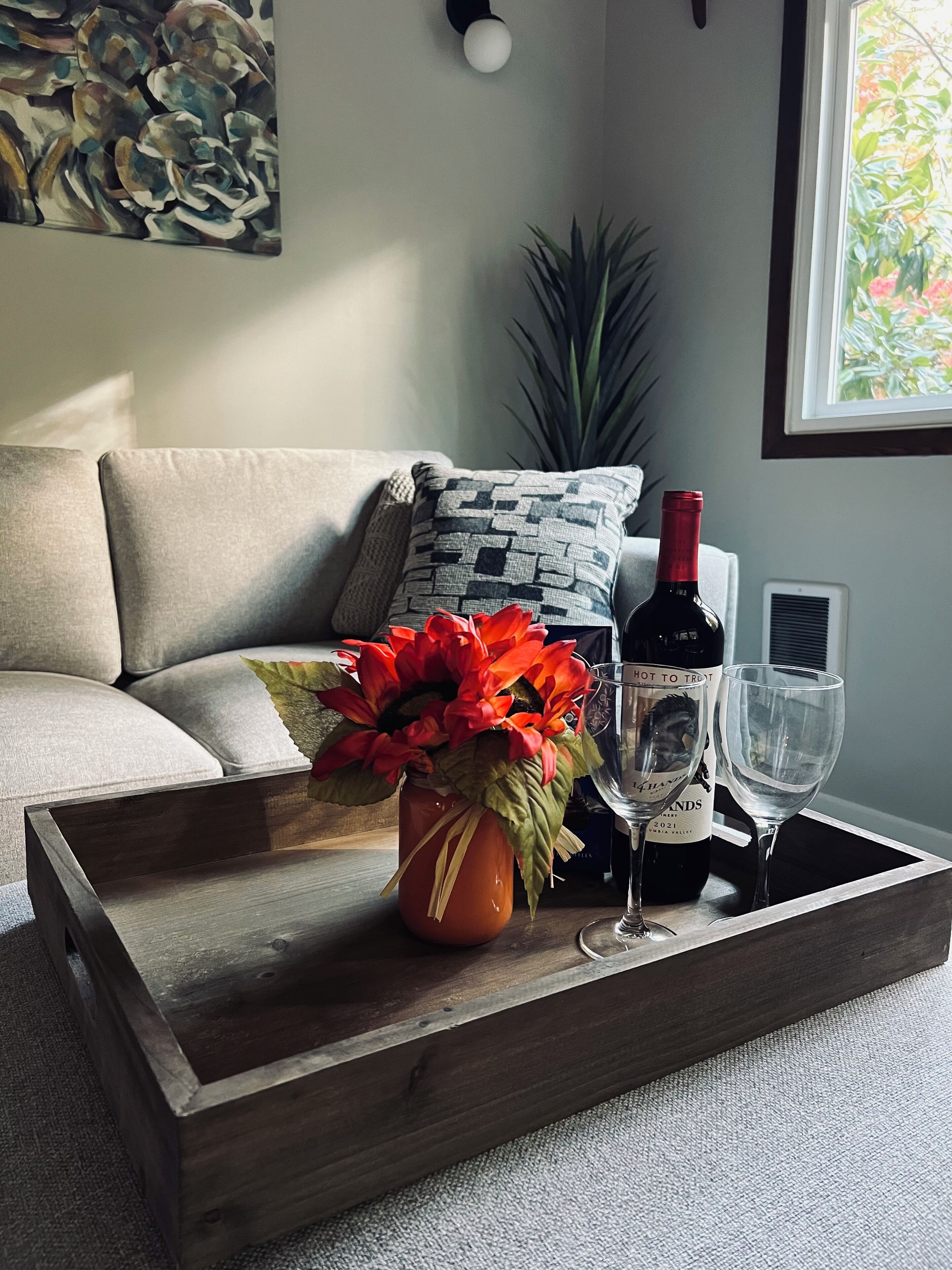 A wooden tray with a wine bottle and glasses on a sofa, a vase with red flowers, and a plant in the background.
