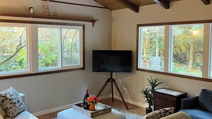 A living room with a wooden ceiling, large windows, a flat-screen TV, a sofa, a coffee table, and a side table with a plant. - Charming Geneva Bungalow (Bellingham)