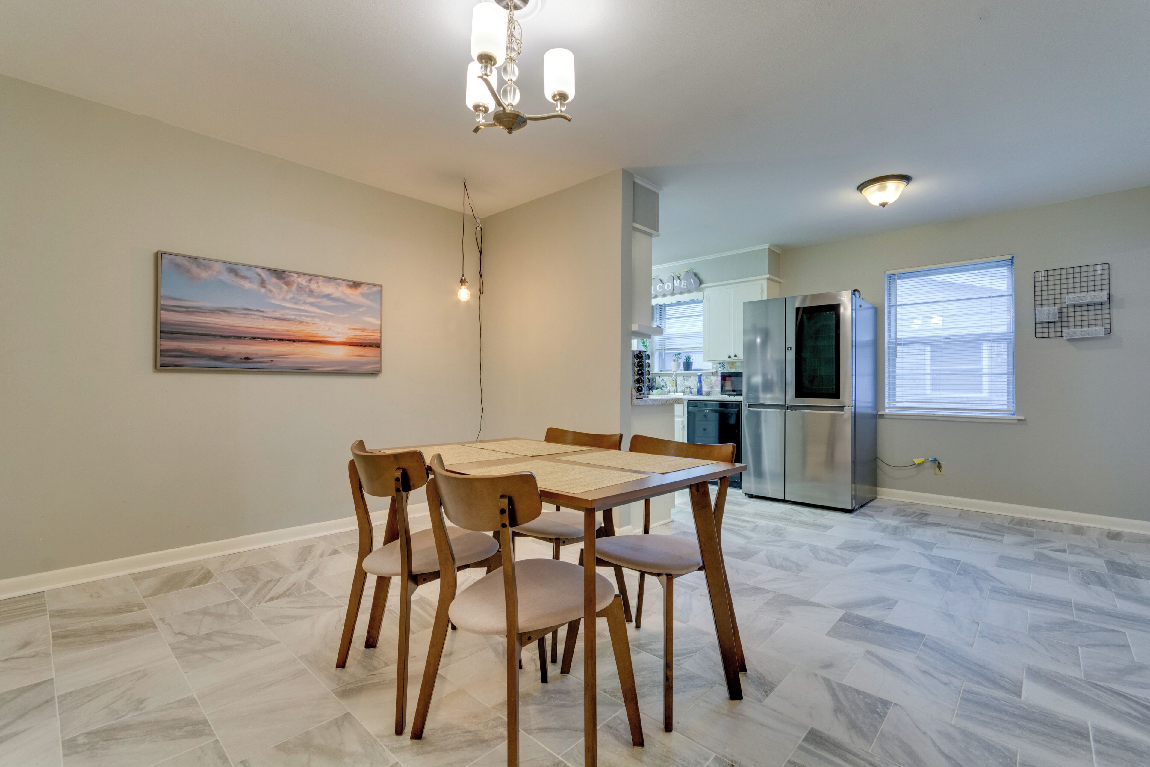 Un coin repas avec une table et des chaises en bois, un lustre, un tableau et, au fond, une cuisine.