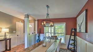 Un coin repas avec une table en bois, des chaises, un lustre et une vue sur l’extérieur par une fenêtre.