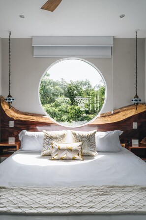Quarto com uma grande janela redonda, cabeceira de madeira e cama com roupa de cama branca e almofadas estampadas.