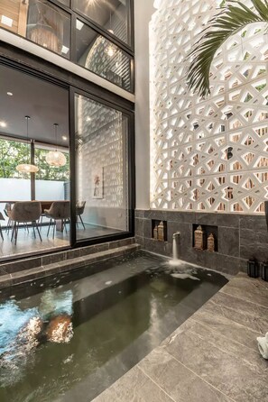 Ein moderner Poolbereich im Innenbereich mit Wasserspiel, einem Essbereich mit Stühlen und Tisch sowie einer Wand mit geometrischen Mustern.