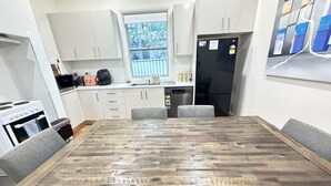 Een eethoek met een houten tafel, gestoffeerde grijze stoelen, witte kasten, een zwarte koelkast en een raam met uitzicht op bomen.