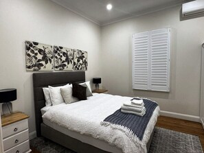 A bedroom with a bed, bedside tables, a wall-mounted air conditioner, and a window with blinds.