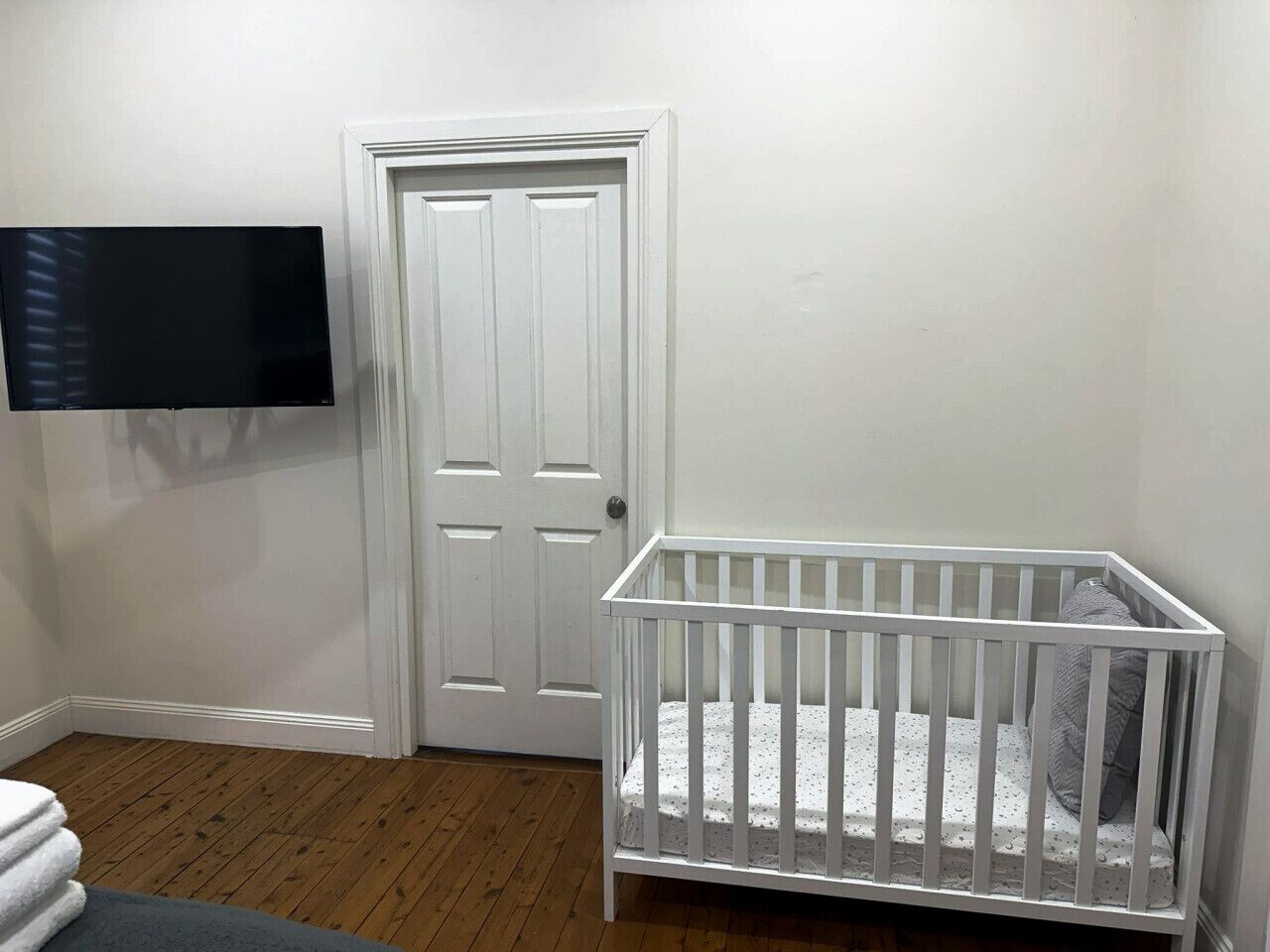 A room with a white door, a white crib, and a flat-screen TV mounted on the wall.