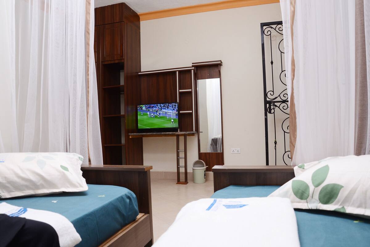 Ein Hotelzimmer mit zwei Betten, einem Fernsehgerät auf einem Tisch, einem Kleiderschrank und einem Badezimmer mit Duschvorhang.