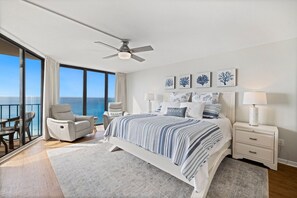A bedroom with a large bed, a ceiling fan, a nightstand, a chair, and a view of the ocean.