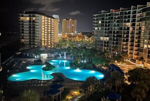 A hotel with a large outdoor swimming pool surrounded by palm trees and modern buildings in the background.