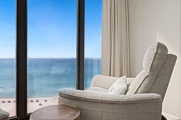 A modern room with a large window overlooking a beach, a grey armchair with cushions, and a small wooden side table.