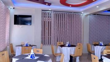 A hotel dining area with round tables, wooden chairs, and a large TV on the wall.
