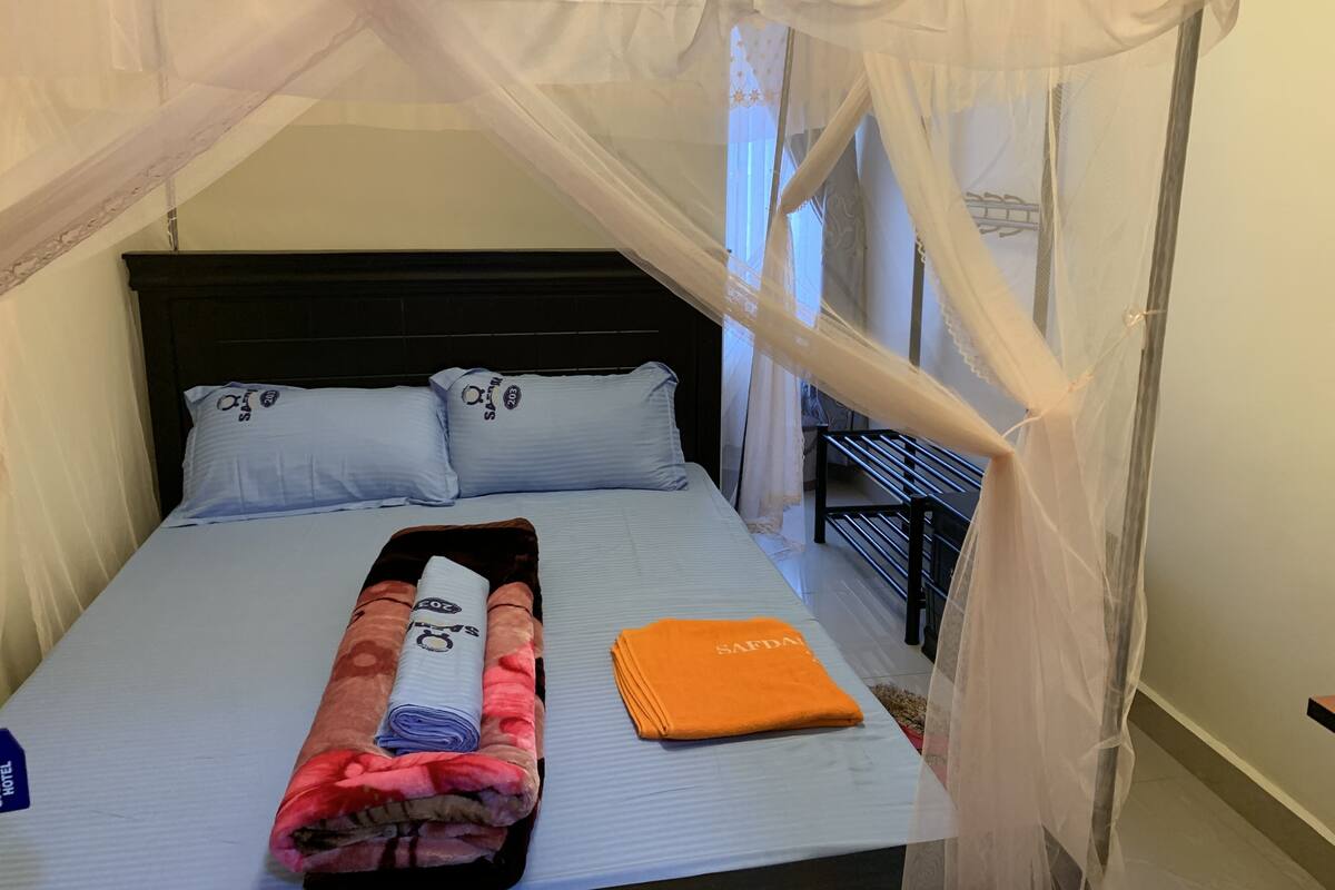Una cama bien hecha con dosel, almohadas, una toalla enrollada y una toalla naranja.