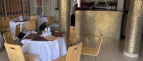 A restaurant with round tables, wooden chairs, and a bar area with bottles.