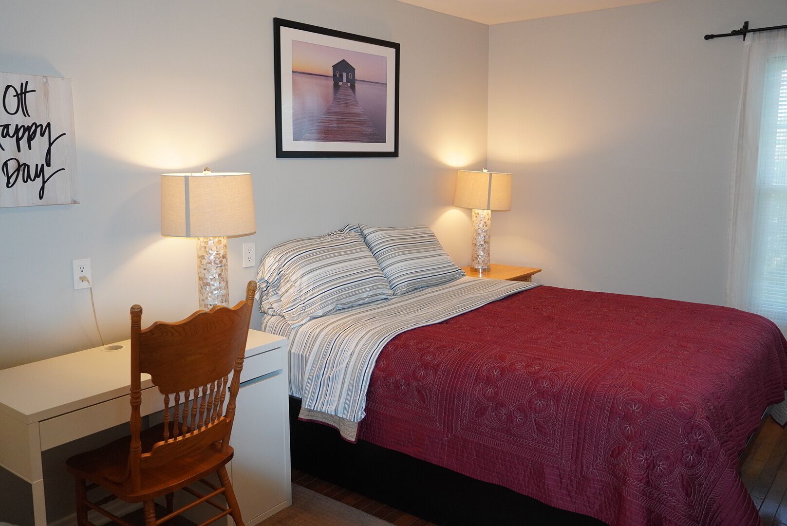 A bedroom with a bed, two bedside lamps, a chair, a desk, and a framed picture on the wall.