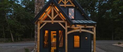 A small, dark-colored cabin with a stone chimney, wooden beams, and a porch with a single door and two windows.