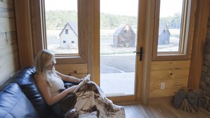 A room with a wooden interior, a black leather couch, and a view of rustic buildings through large glass doors.