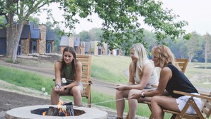 Tres personas sentadas alrededor de una fogata en un entorno al aire libre, con braseros de piedra y sillas de madera.