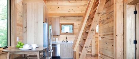 A wooden cabin interior with a dining area, kitchen, and a staircase leading to an upper level.