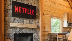 Ein gemütliches Wohnzimmer mit Kamin aus Stein, einem Netflix-Schild, Holzbalken und Blick ins Freie.