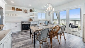 A spacious kitchen with white cabinetry, a dining area with wicker chairs, and a view of the ocean through large windows.