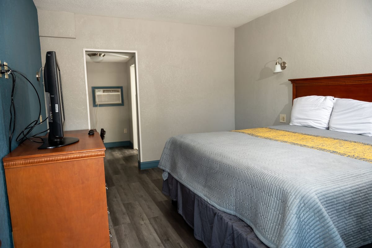 A hotel room with a bed, a wooden dresser, a television on the dresser, and a door leading to another room.