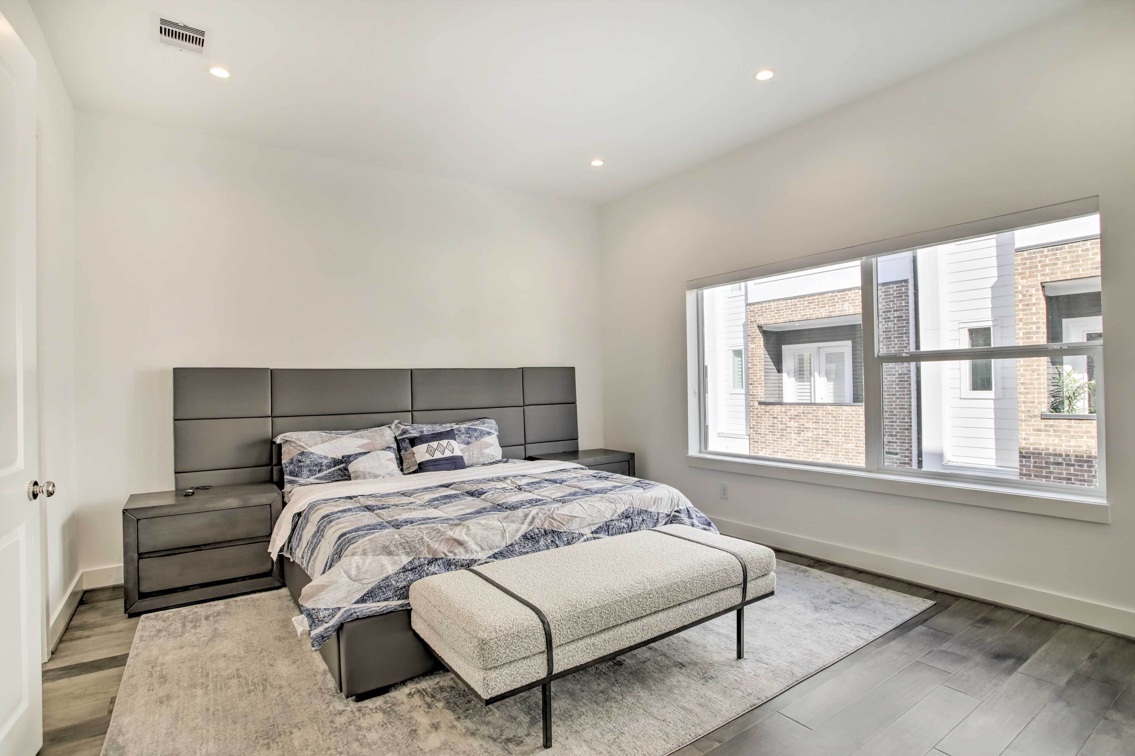 A bedroom with a large bed, a nightstand, a bench, and a window with a view of a brick building.