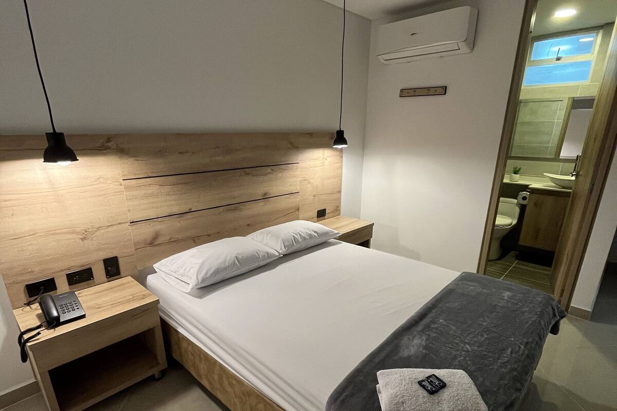 Une chambre moderne avec une tête de lit en bois, une table de chevet avec un téléphone, une lampe de chevet et un lit recouvert d’une couverture et de coussins gris.