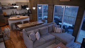 Eine moderne Küche mit einer Holzinsel, ein Essbereich mit großem Tisch und Stühlen, ein graues Sofa und ein Ausblick ins Freie durch große Fenster.