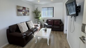 Una sala de estar con dos sofás de cuero marrón oscuro, una mesa de centro blanca, un televisor de pantalla plana grande montado en la pared y una ventana con persianas.