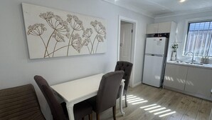 Un comedor con una mesa blanca y cuatro sillas, una cocina con refrigerador y armarios, y una obra de arte floral colgada en la pared.