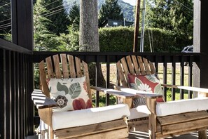 Een houten terras met twee Adirondack-stoelen, waarvan er een een bloemenkussen heeft en de andere een bladerenpatroon kussen.