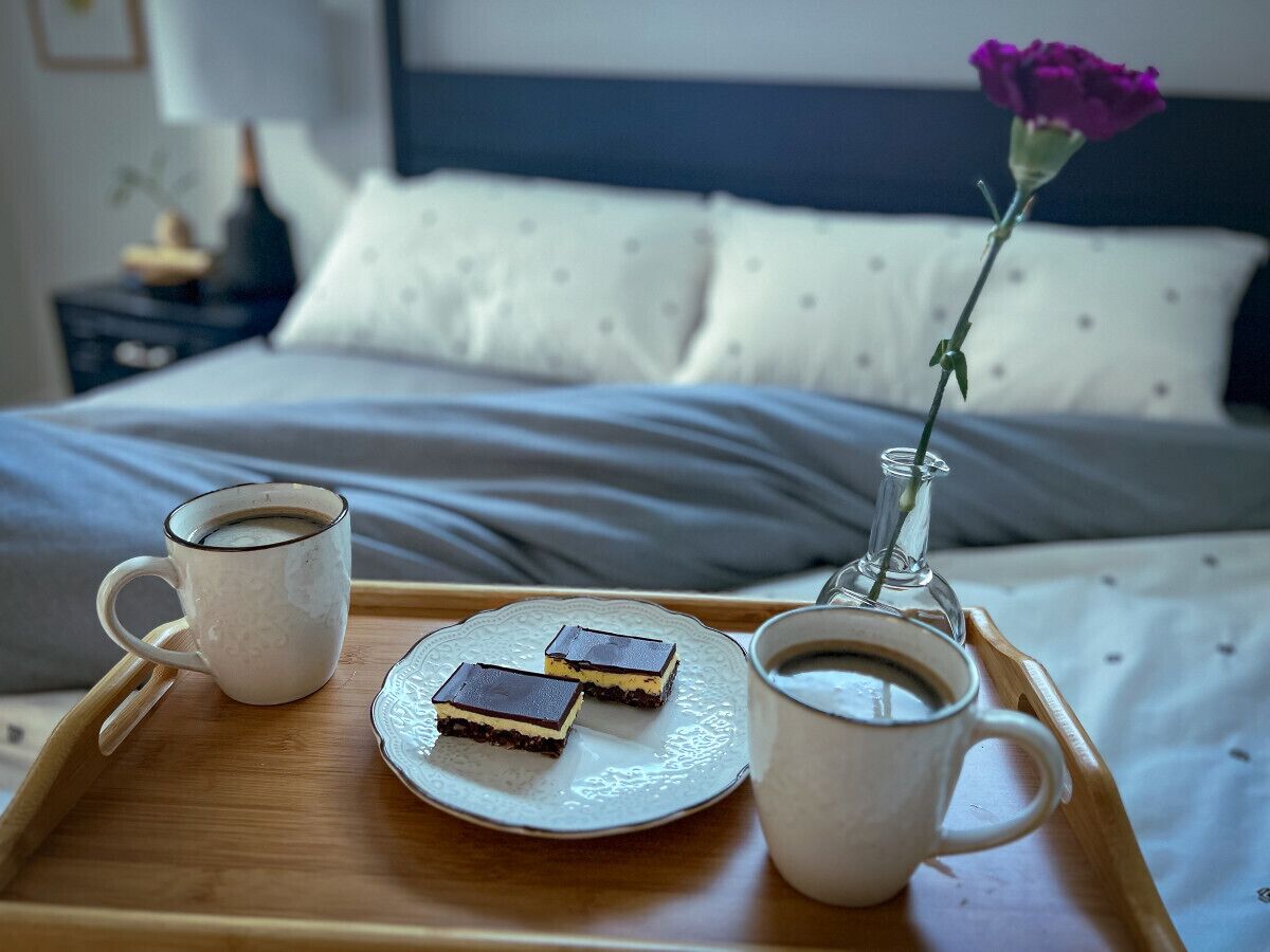 A tray with two cups of coffee, a plate with two desserts, and a vase with a single flower on a bed.