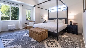 A four-poster canopy bed with a white bedspread, a bedside table with a lamp, and a small side table with books. - Luxury 4BR in the Foothills · Lake & Trails · Sleeps 8 (Sedro-Woolley)