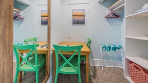A room with a wooden table and two green chairs, a white door with a sunset painting, and a white shelf with a pink basket.