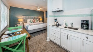 Een kleine hotelkamer met een bed, nachtkastje, lamp en een kitchenette met een spoelbak en koffiezetapparaat.