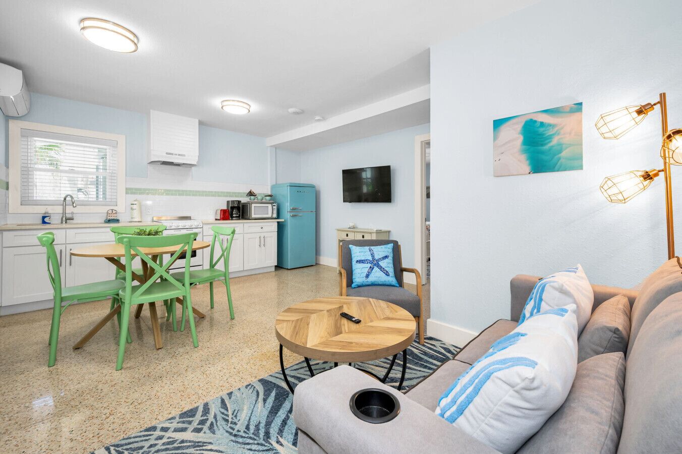 A compact living space with a kitchenette, a dining area with green chairs, a sofa with blue pillows, and a TV mounted on the wall.