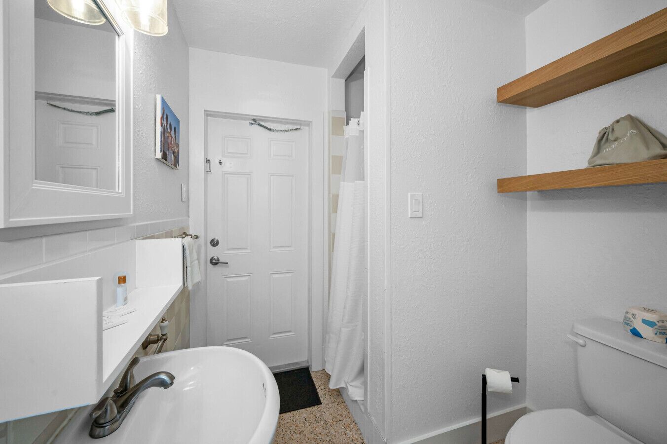 Een badkamer met een wit wastafel, spiegel en toilet. Er is een douchegordijn en een houten plank boven het toilet.