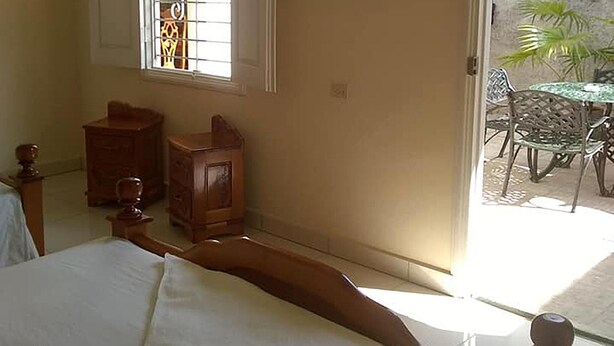 Ein Schlafzimmer mit einem großen Bett, einem Fenster mit Gitterstäben, einer kleinen Kommode und Blick auf eine Außenveranda mit Stühlen und einem Tisch.