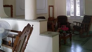 Ein geräumiges Wohnzimmer mit hoher Decke, einer Kandelaber, einem großen Fenster mit Jalousien und mehreren Holzstühlen sowie einem Tisch.