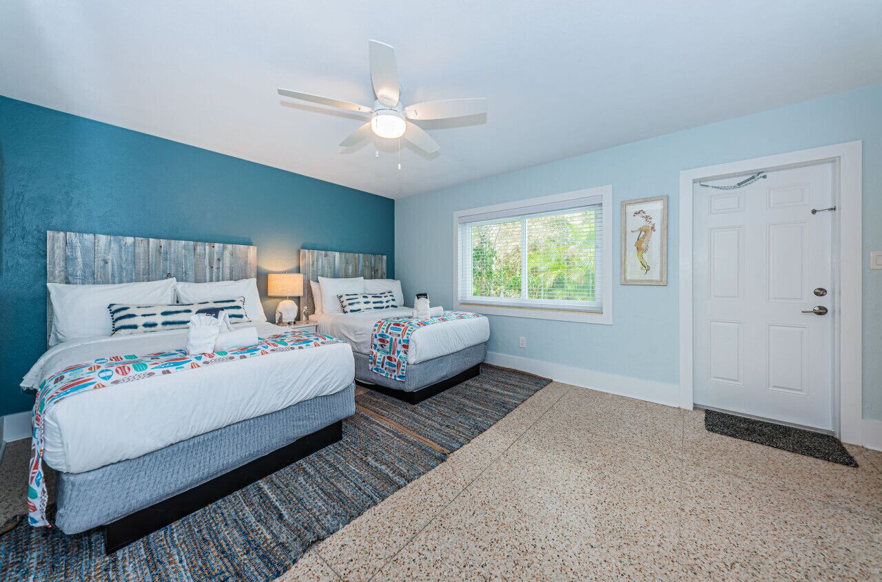 A bedroom with two beds, a ceiling fan, a window with a view, and a white door.