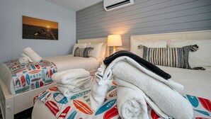 A hotel room with two beds, a wall-mounted air conditioner, a painting of a sunset, and a lamp.