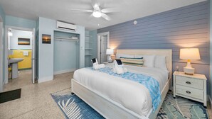 A hotel room with a bed, bedside tables, a ceiling fan, an air conditioning unit, a bathroom with a sink and mirror, and a view of the beach.