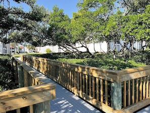 A wooden boardwalk with a metal grid floor, surrounded by lush greenery and trees. - Runaway Bay 239-Bayfront view - Pool, tennis, pickleball-Resort style community (Bradenton Beach)